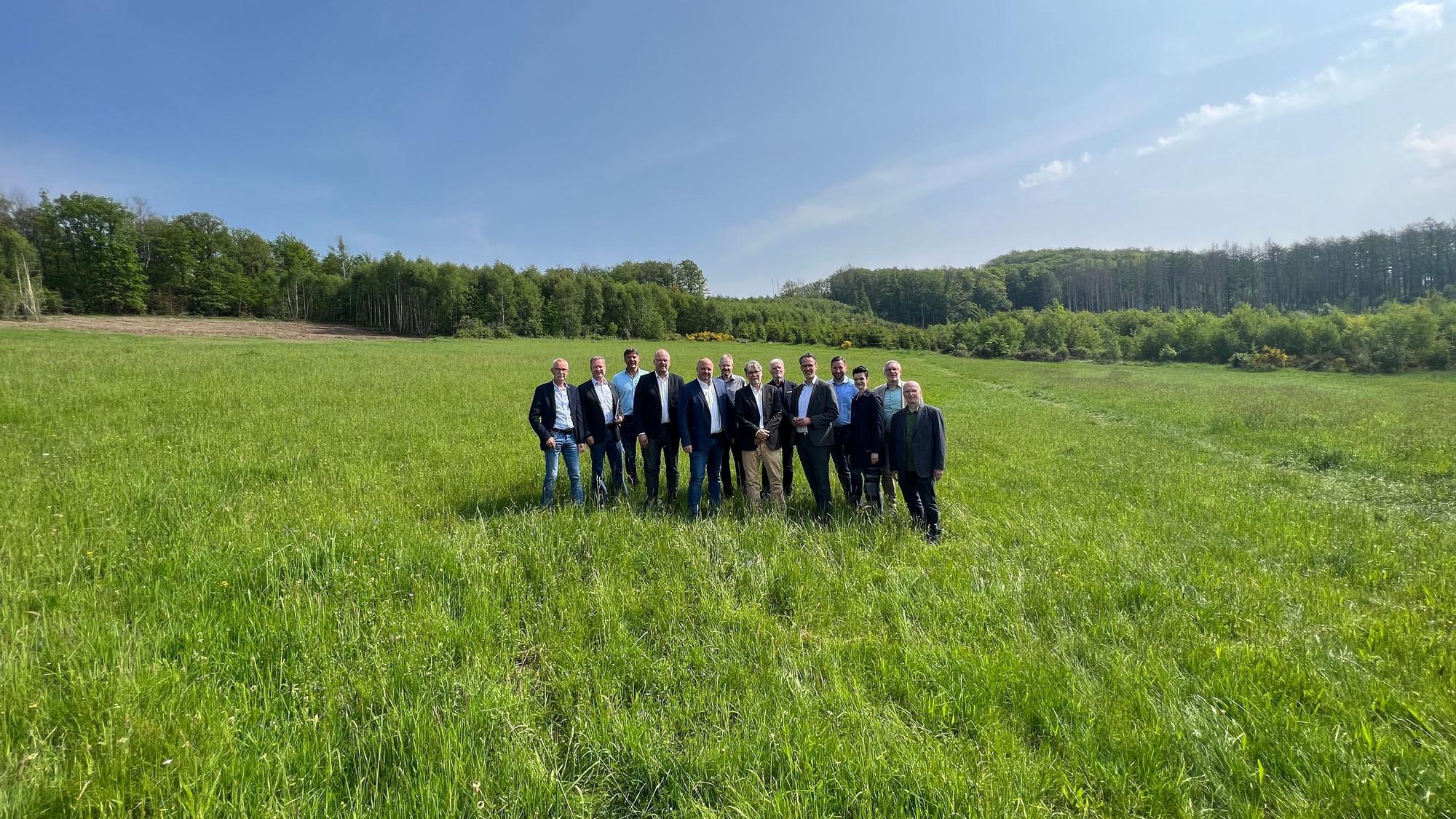 Eine große, grüne Wiese mit Bäumen im Hintergrund und strahlend blauem Himmel. In der Mitte steht eine Gruppe aus 13 Personen.