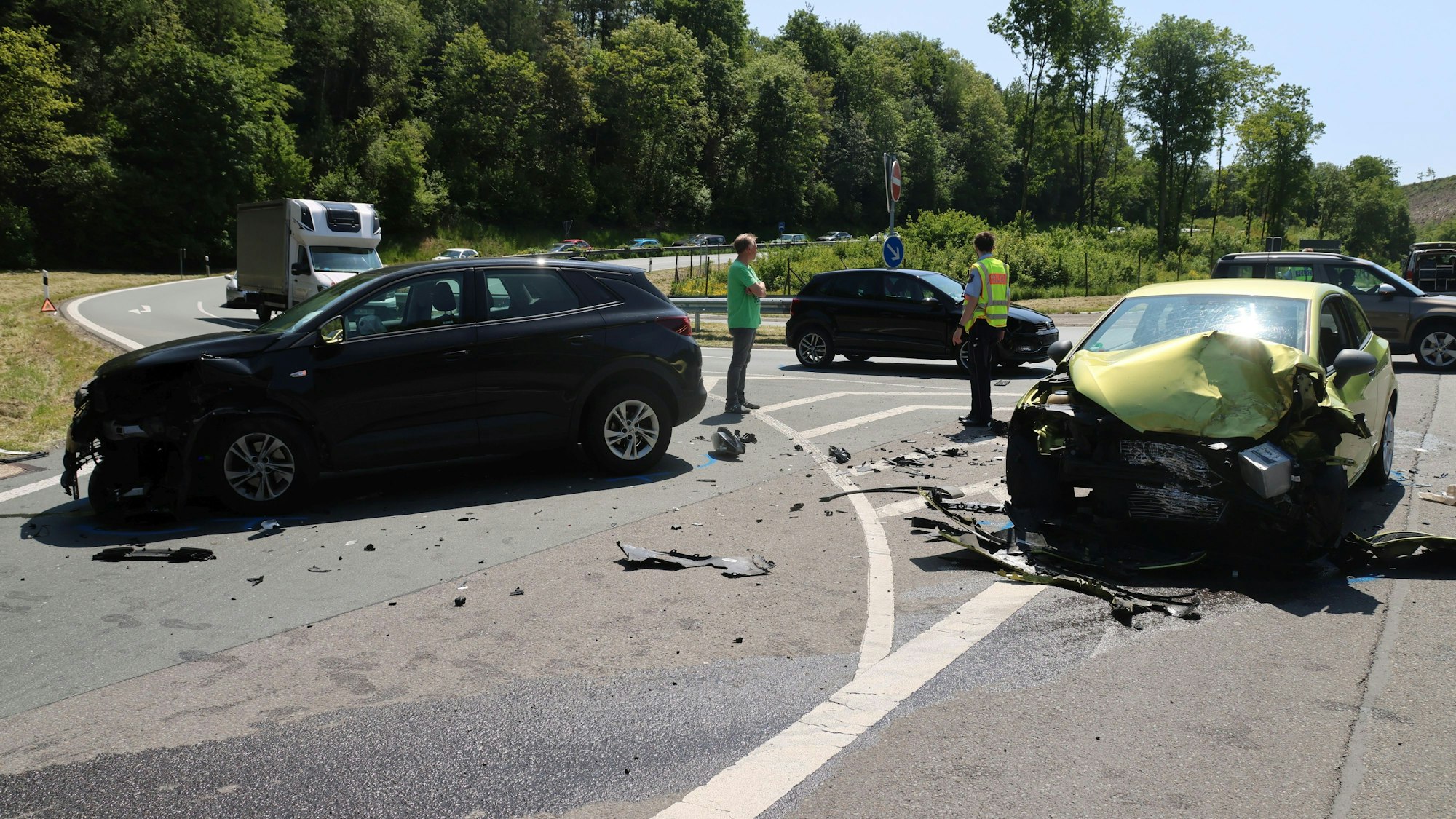 Drei Autos stehen schwer beschädigt auf der Straße. Zwischen Ihnen eine Person in grünem T-Shirt und ein Polizist.
