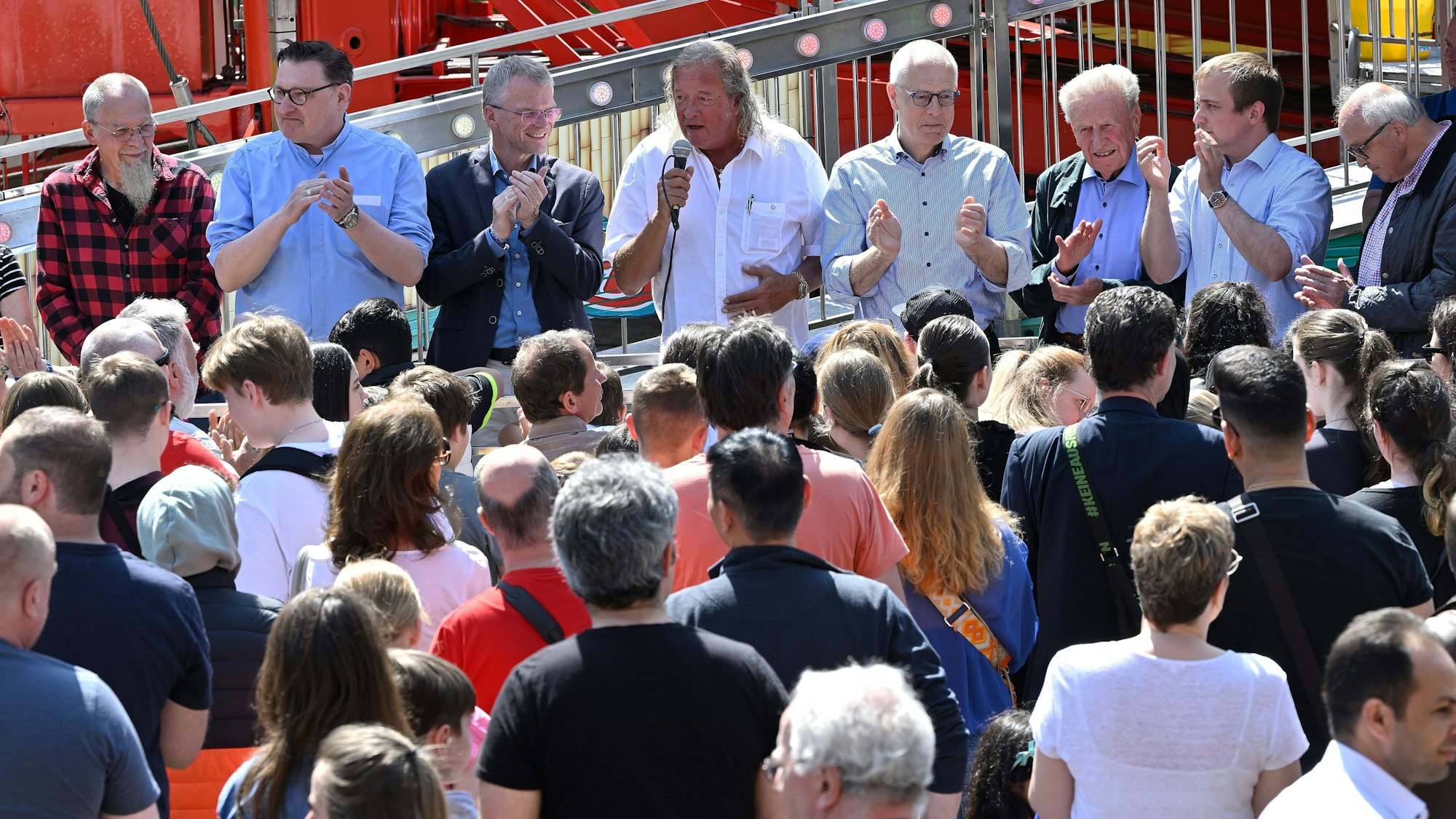 Einige Menschen stehen auf einem Kirmesfahrgeschäft und applaudieren. Einer hält ein Mikro in der Hand. Vor dem Fahrgeschäft stehen zahlreiche Menschen, die zu den Menschen auf dem Fahrgeschäft schauen.
