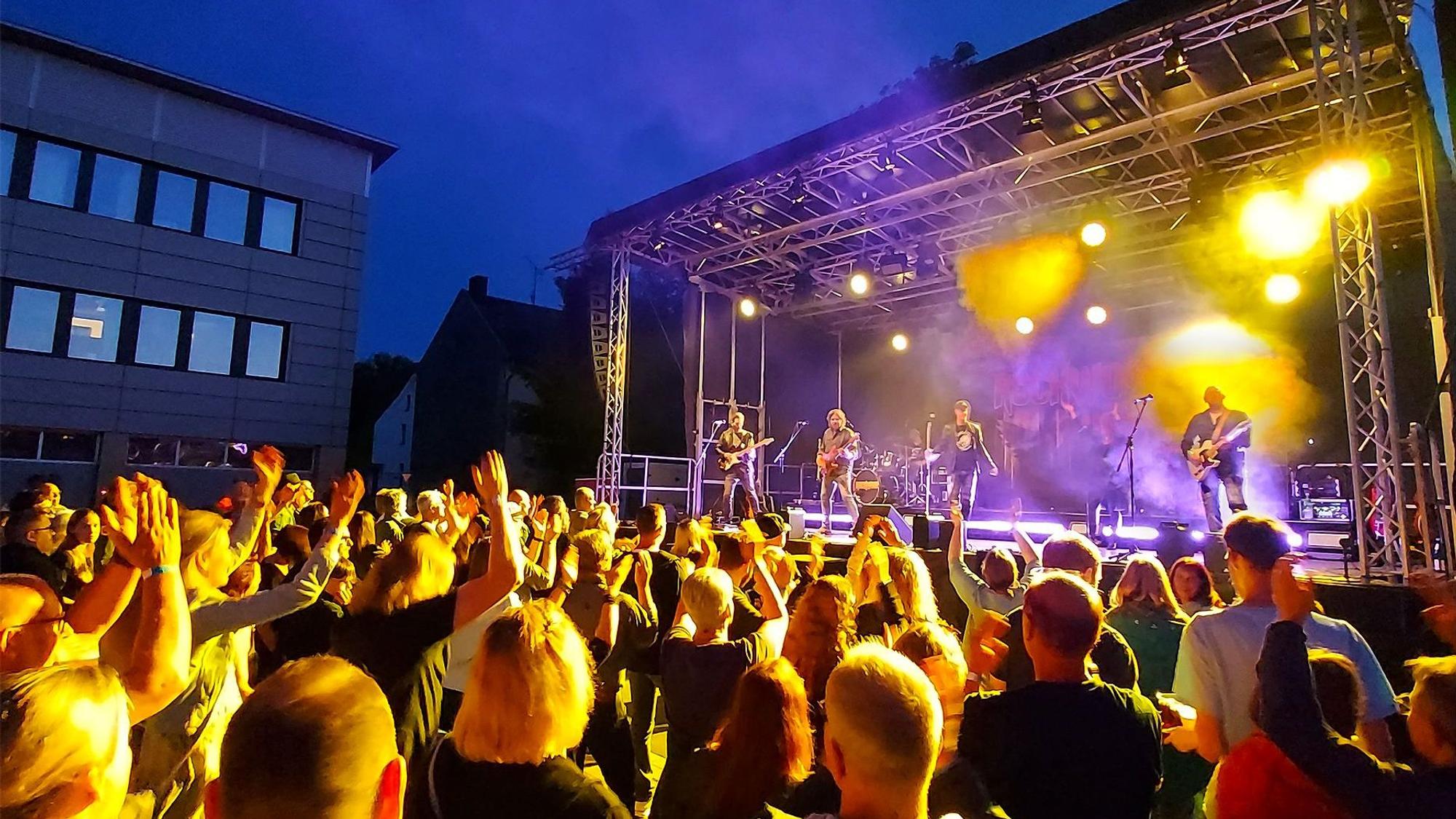 Musikfans stehen vor einer Bühne, auf der Band spielt. Sie werden von den bunten Scheinwerfern angestrahlt.