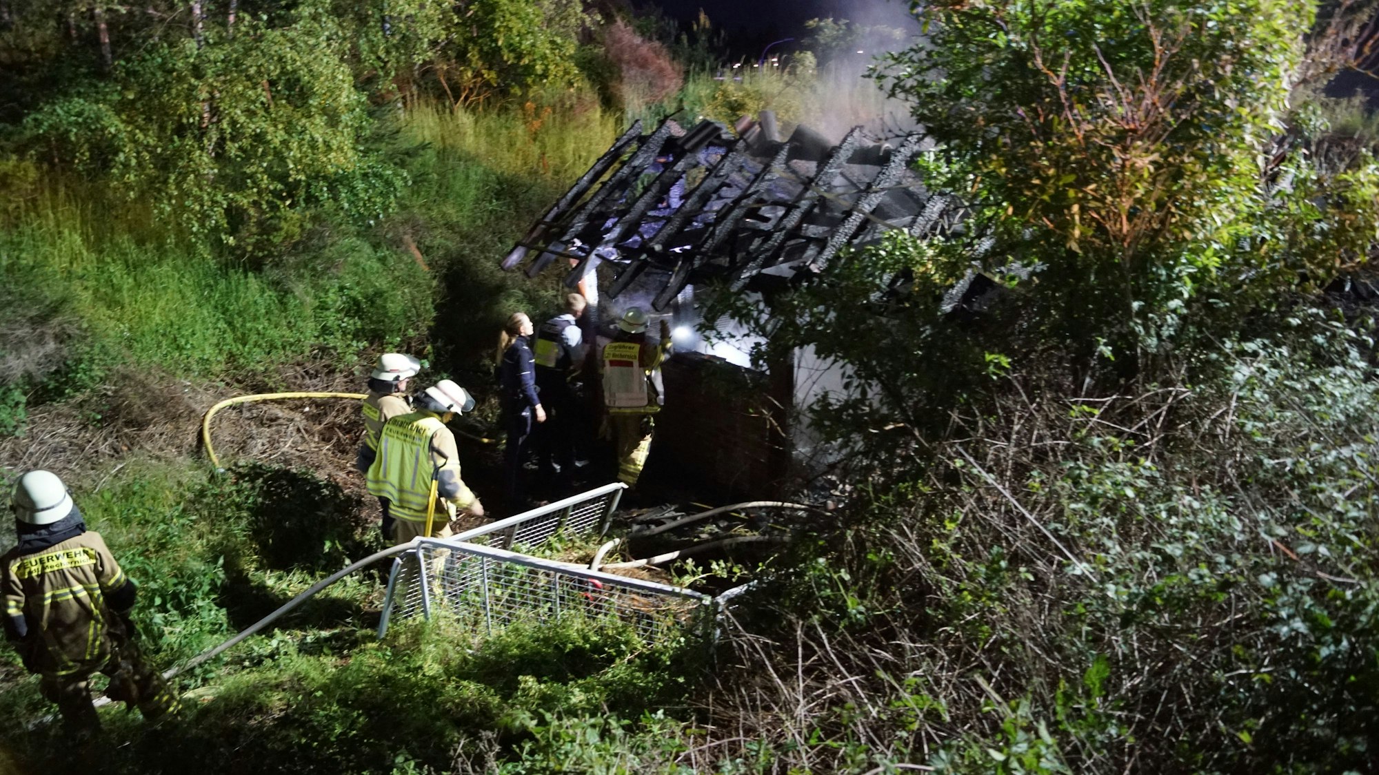 Einige Feuerwehrleute und Polizisten stehen an der ausgebrannten Grillhütte in Mechernich.