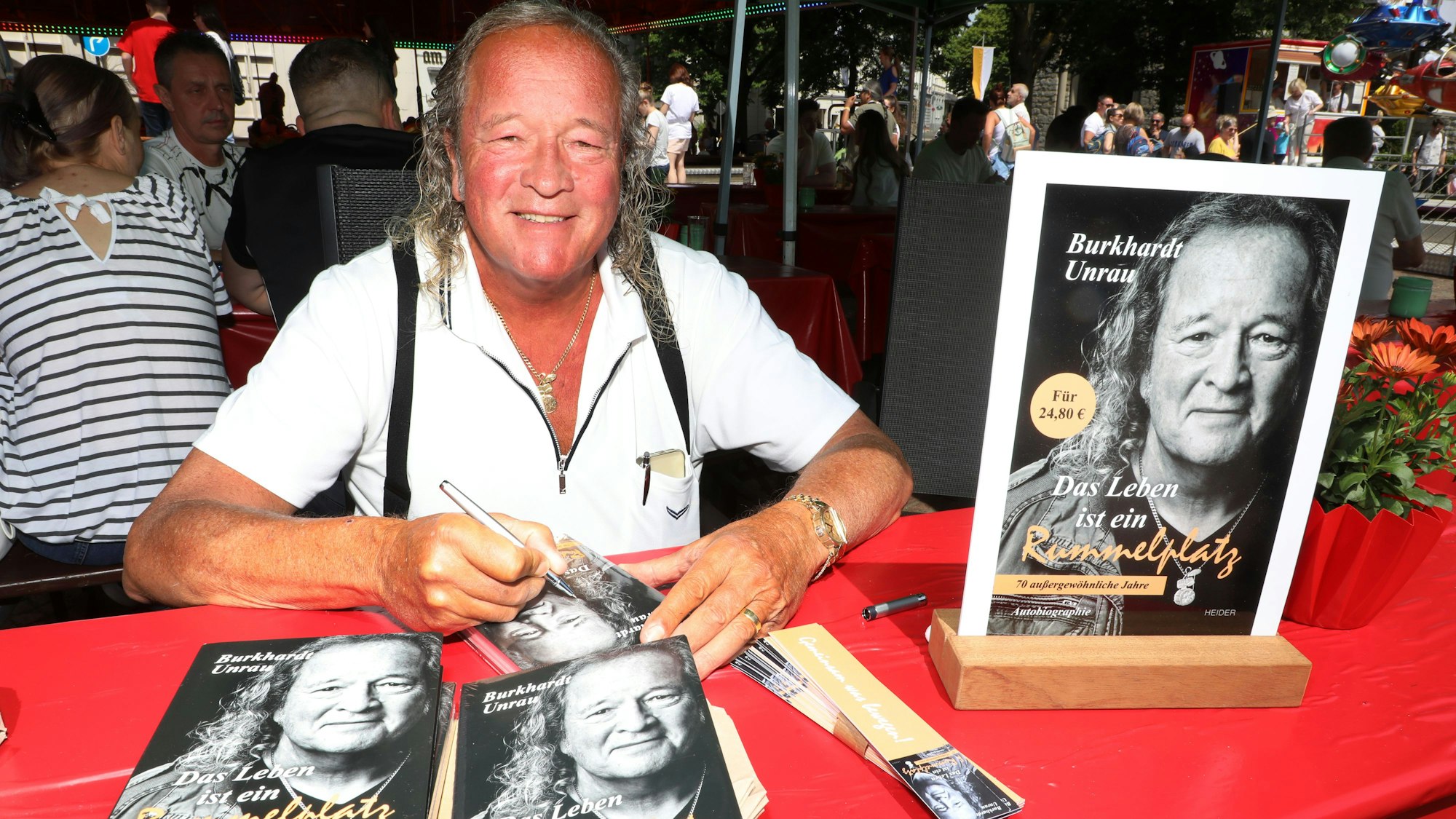 Kirmesmacher Burkhardt Unrau sitzt an einem Tisch mit roter Tischdecke und signiert Bücher seiner Autobiographie „Das Leben ist ein Rummelplatz“.