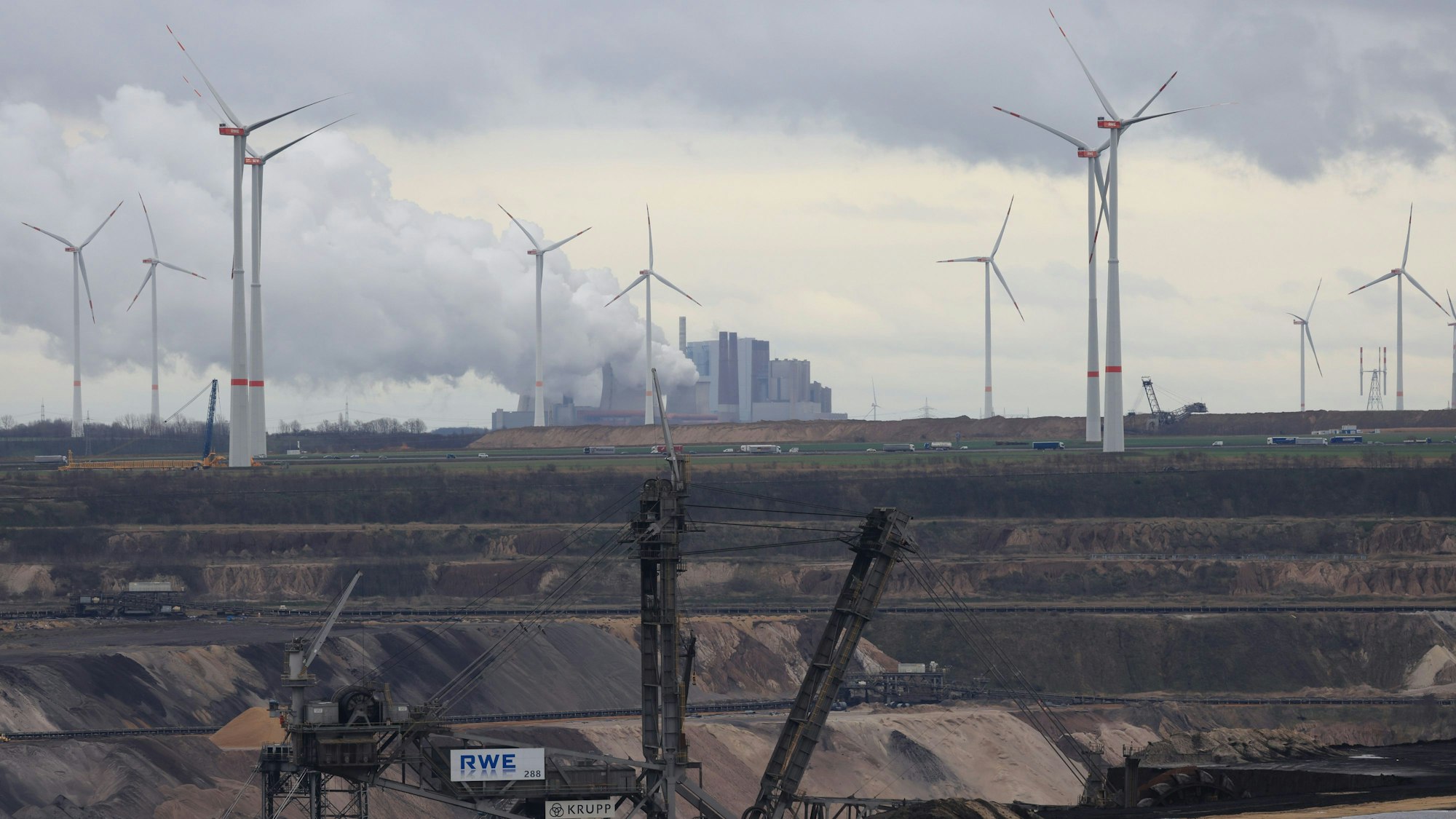 Der Blick auf den Braunkohletagebau Garzweiler II. zeigt schwere Maschinen der RWE im Fordergrund, die Rauch ausstoßen und Windräder im Hintergrund vor einem bewölkten Himmel.