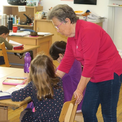 Die Seniorin steht neben einer Schülerin und erklärt ihr eine Aufgabe,