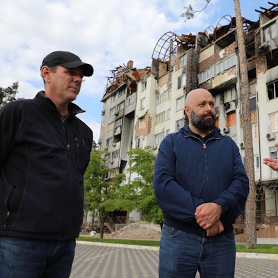 Drei MEnschen stehen sprechend vor einem zerbombten und ausgebrannten Wohn- und Geschäftshaus bei Butscha.