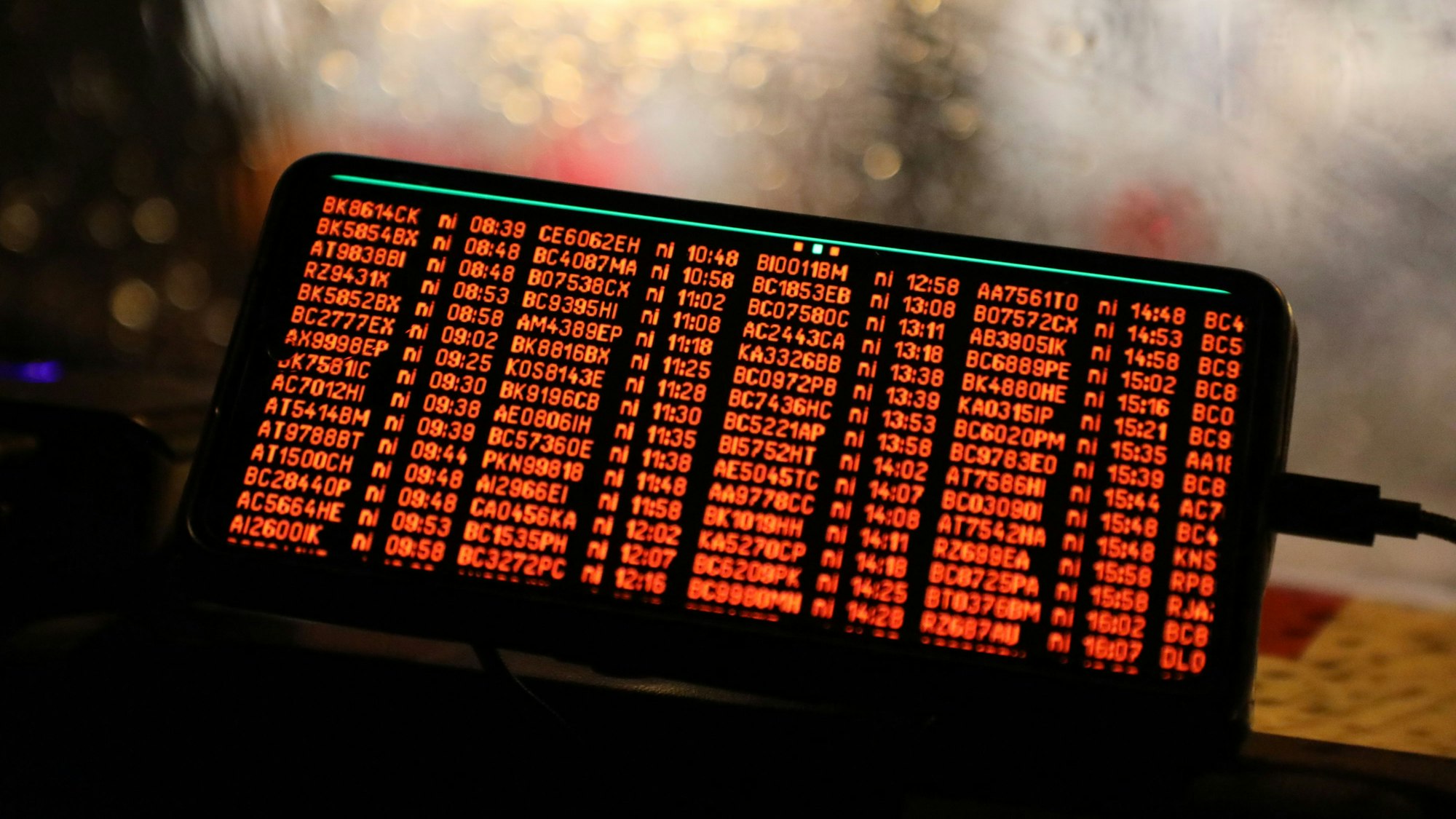 Ein Handy mit roten Ziffern und Buchstaben ist auf dem Armaraturenbrett eines Autos zu sehen. Regentropfen laufen die Windschutzscheibe dahinter herunter.