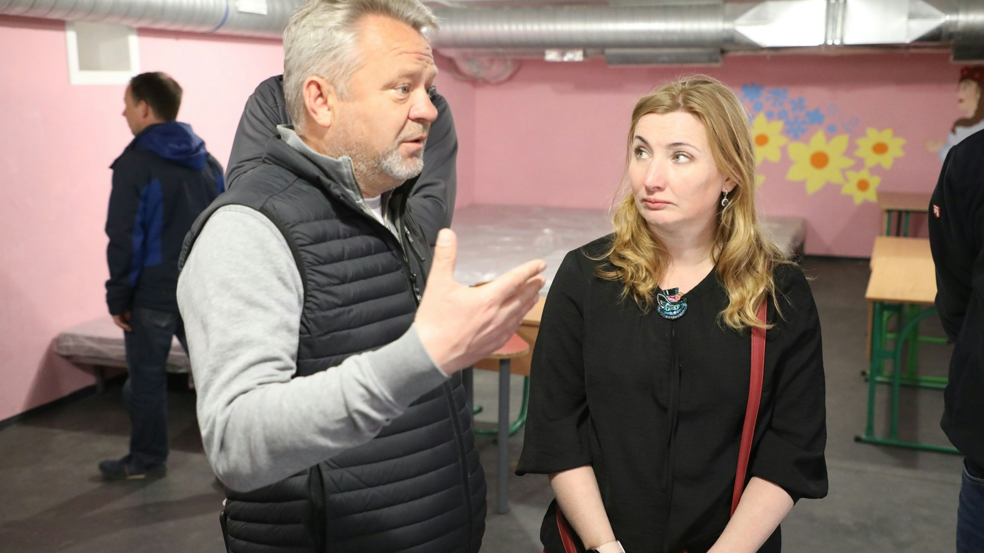 Ein Mann und eine Frau stehen in einem Bunker, an dessen Wände Blumen in Geld und Blau gemalt sind. Im Hintergrund stehen Betten und Schultische.