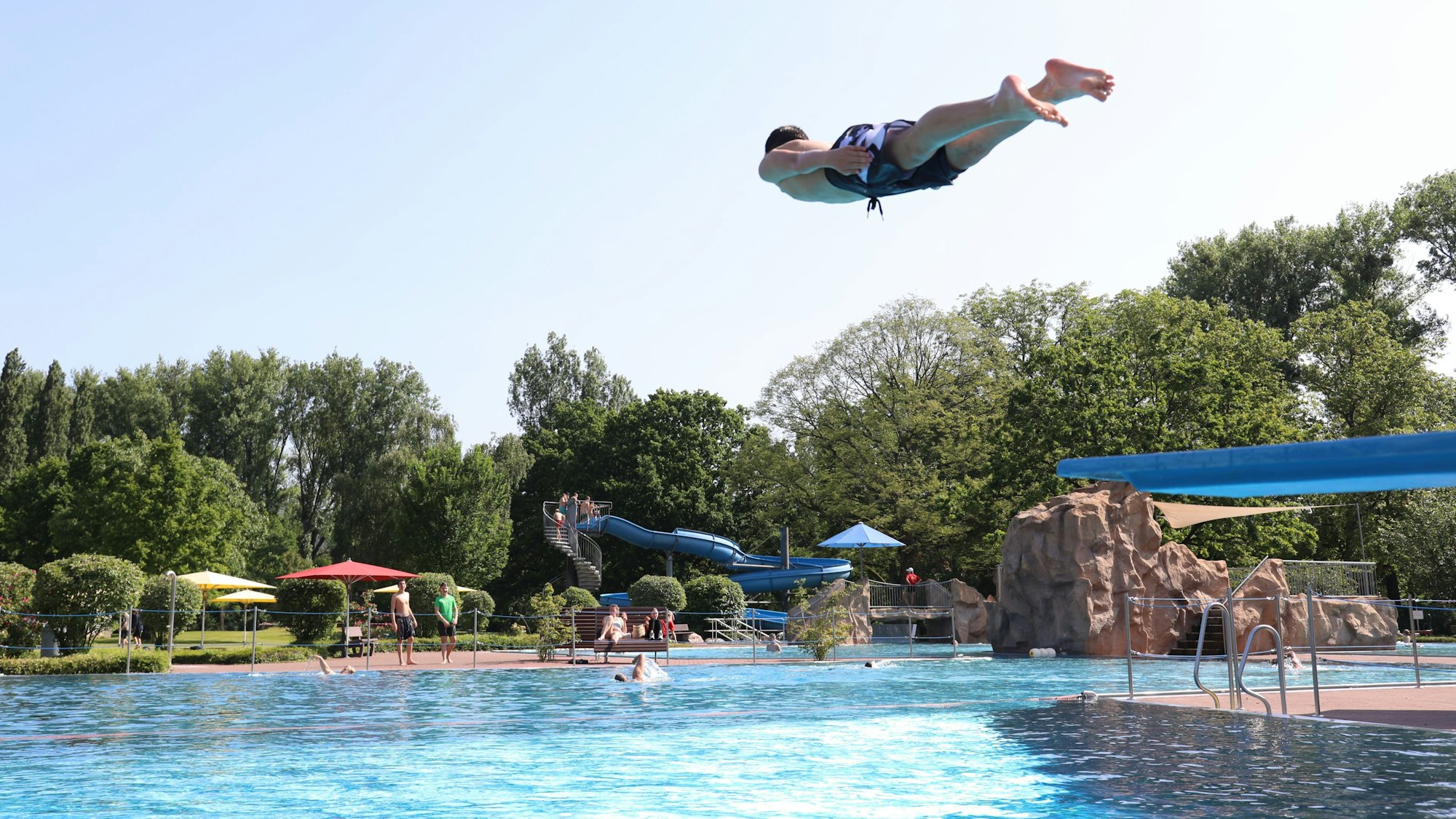 Ein Jugendlicher springt von einem Sprungbrett ins Freibad, im Hintergrund eine große blaue Wasserrutsche.