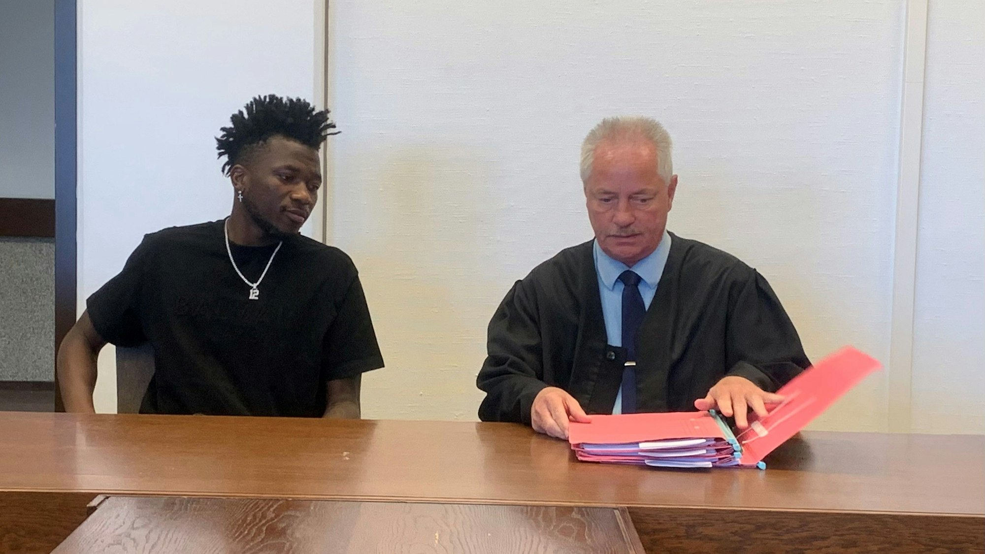 Der Verteidiger von Bayer Leverkusen, Edmond Tapsoba, sitzt mit einer Kette neben einem Mann, der einen Ordner aufschlägt.