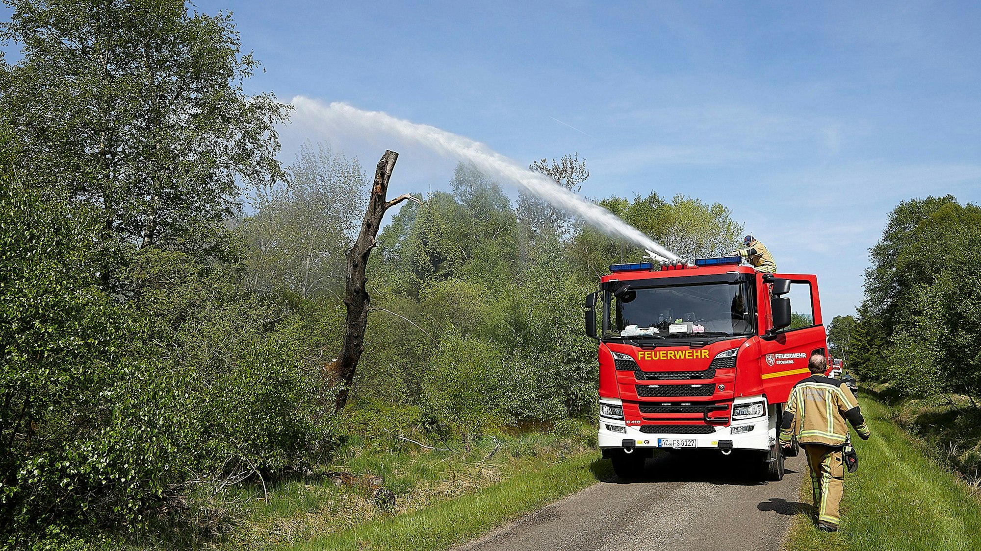Ein Löschfahrzeug spritzt Wasser in die Büsche, ein Feuerwehrmann steht vor dem Auto.