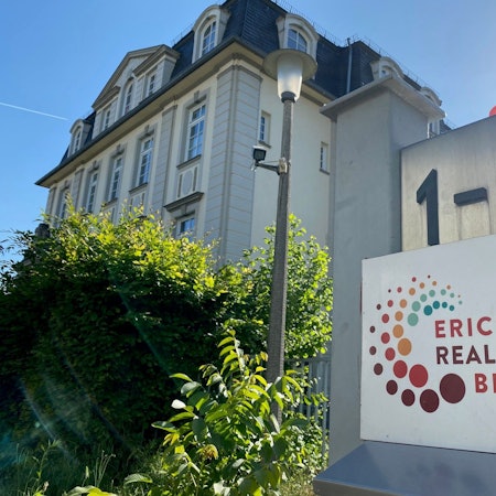 Die Erich-Kästner-Realschule in Brühl ist abgebildet. Die Hausnummer, das Logo der Realschule sowie das Schulgebäude sind zu sehen.
