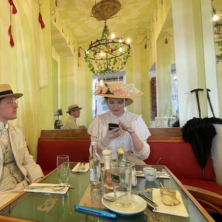 Ein Mann mit Hut und hellem Anzug mit Weste und Krawatte und eine Frau mit einem mit Blumen besetztem Hut auf dem Kopf und einem rüschenbesetzten weißen Kleid sitzen an einem Tisch in einem Café.