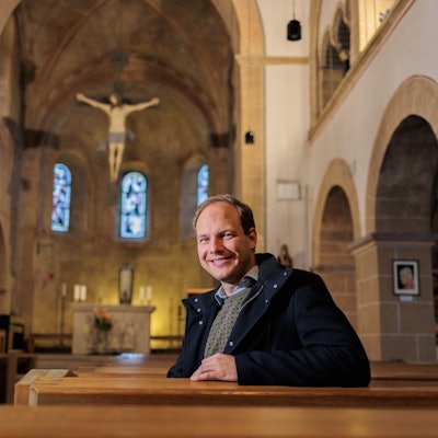 Der Priesteramtsanwärter Sebastian Appolt sitzt auf einer Bank in der Kirche St. Gertrudis und lächelt in die Kamera.