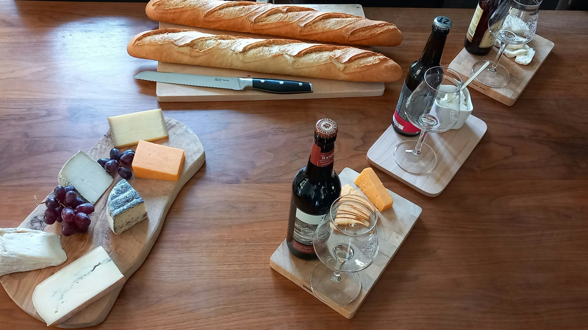 Auf einem Tisch sind verschiedene Flaschen Bier mit Gläsern, eine Käseplatte mit Trauben und zwei Baguetts mit einem Messer angerichtet.