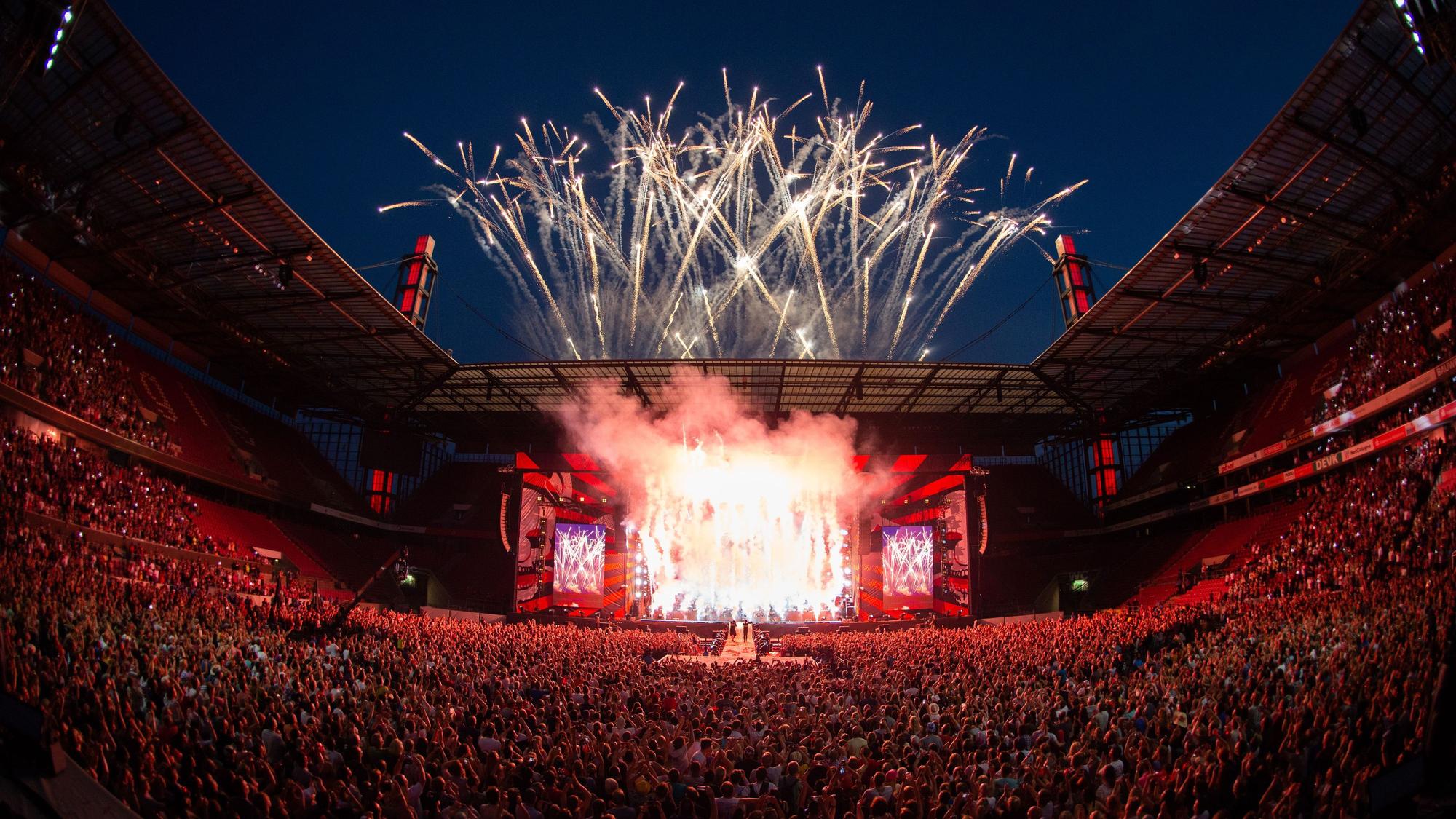 Über dem Rheinenergiestadion explodieren Feuerwerkskörper, die das Publikum im Stadion in rotes Licht tauchen.