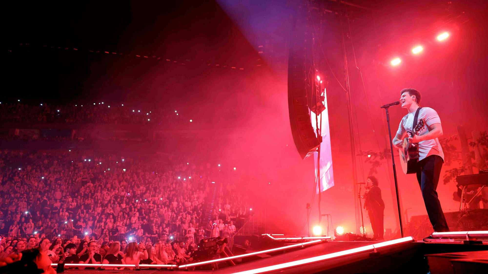 Wincent Weiss spielt auf einer Bühne vor Publikum mit teils leuchtenden Handybildschirmen Gitarre und singt in ein Mikrophon.