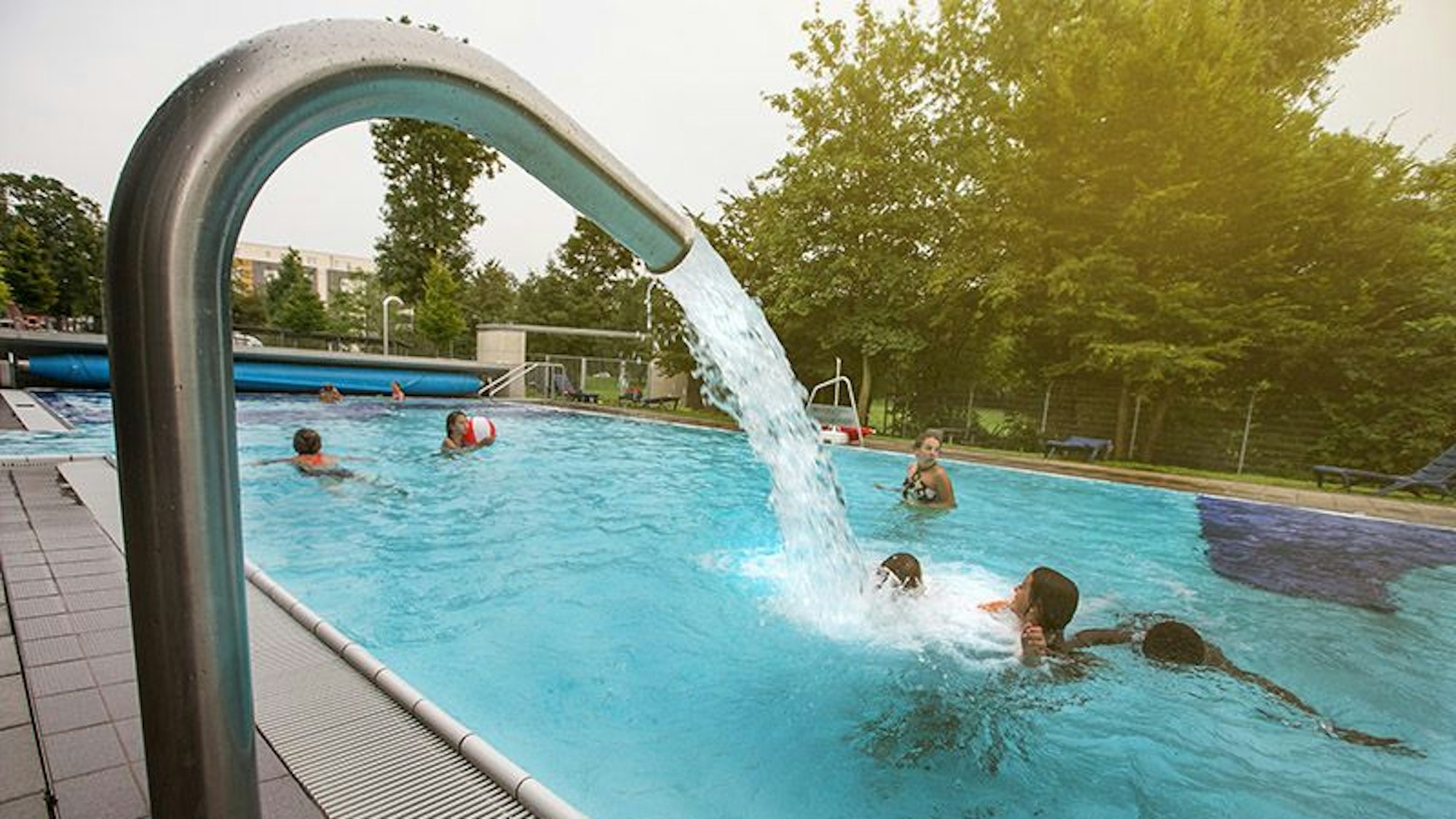 Bei der Aufnahme des Vierjahreszeitenbeckens des Ossendorfbads schießt Wasser aus einem Rohr am Seitenrand und begießt Badegäste im Becken.