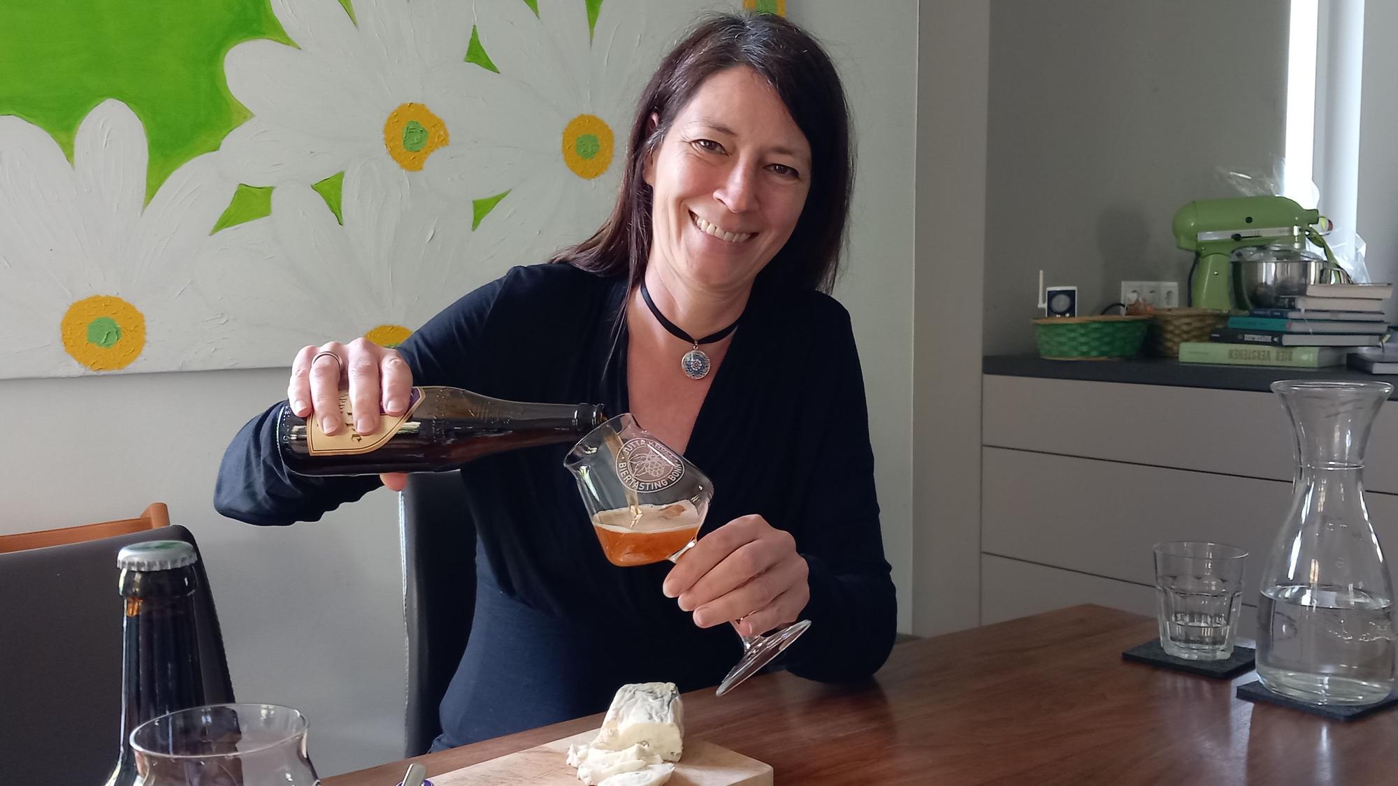 Biersommelière Jutta Knoll aus Bonn-Mehlem schenkt grinsend Bier in ein Glas ein, während sie am Tisch in einer Küche sitzt.