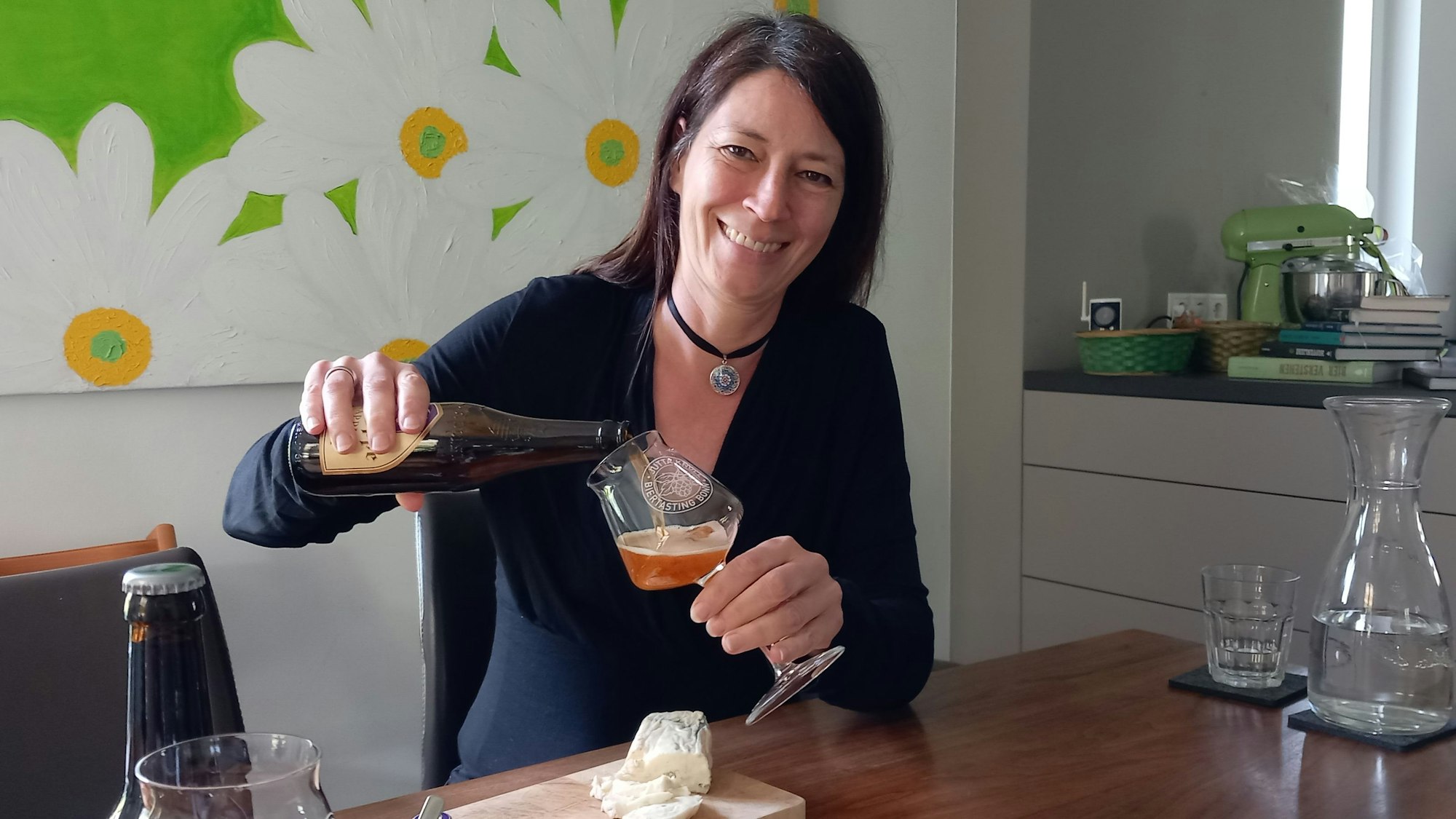 Biersommelière Jutta Knoll aus Bonn-Mehlem schenkt grinsend Bier in ein Glas ein, während sie am Tisch in einer Küche sitzt.