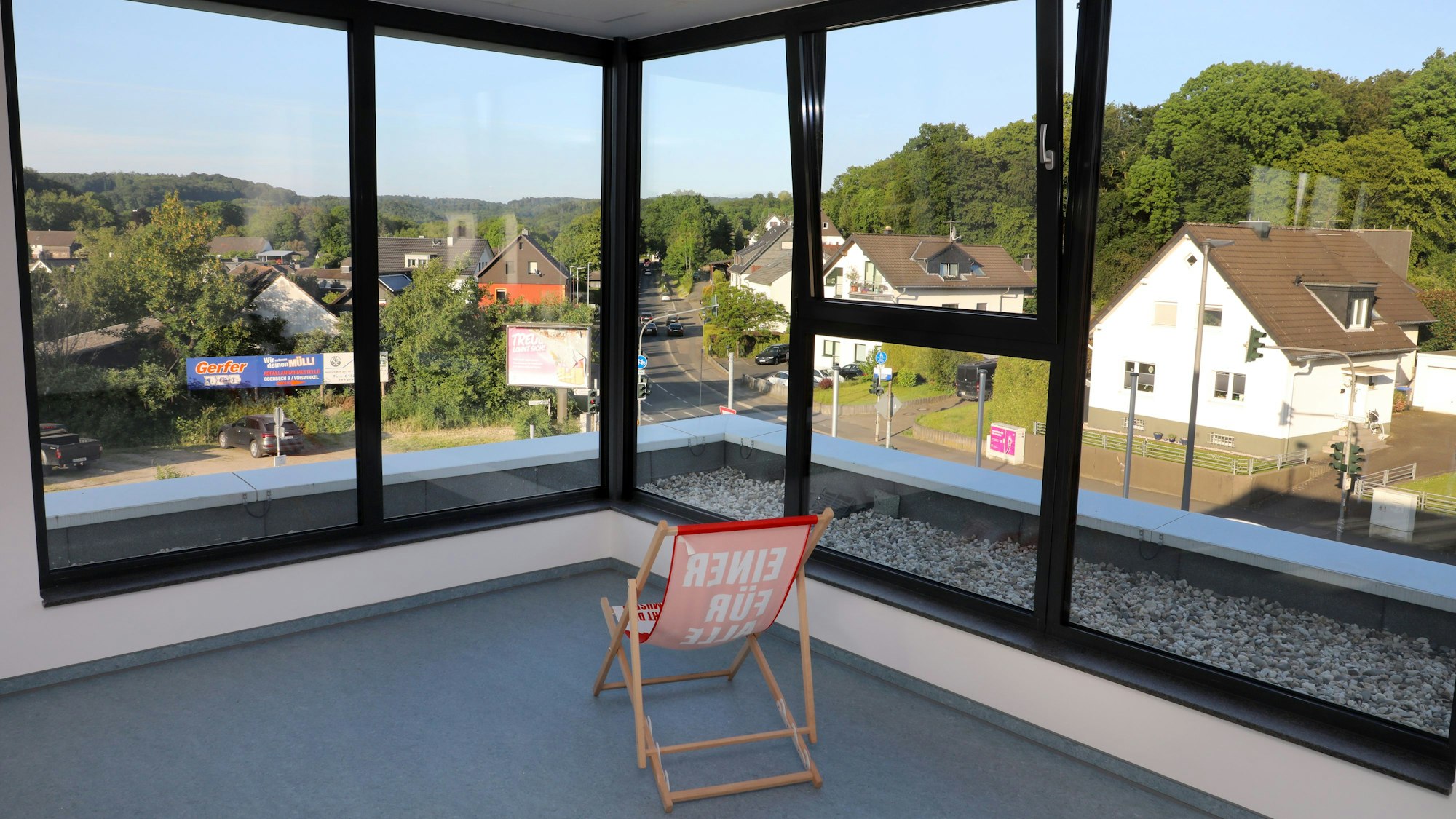 Vor großen Panoramafenstern mit einem reizvollen Ausblick auf Huder, Straßen udn Bäume steht ein Liegestuhl in einem Raum.