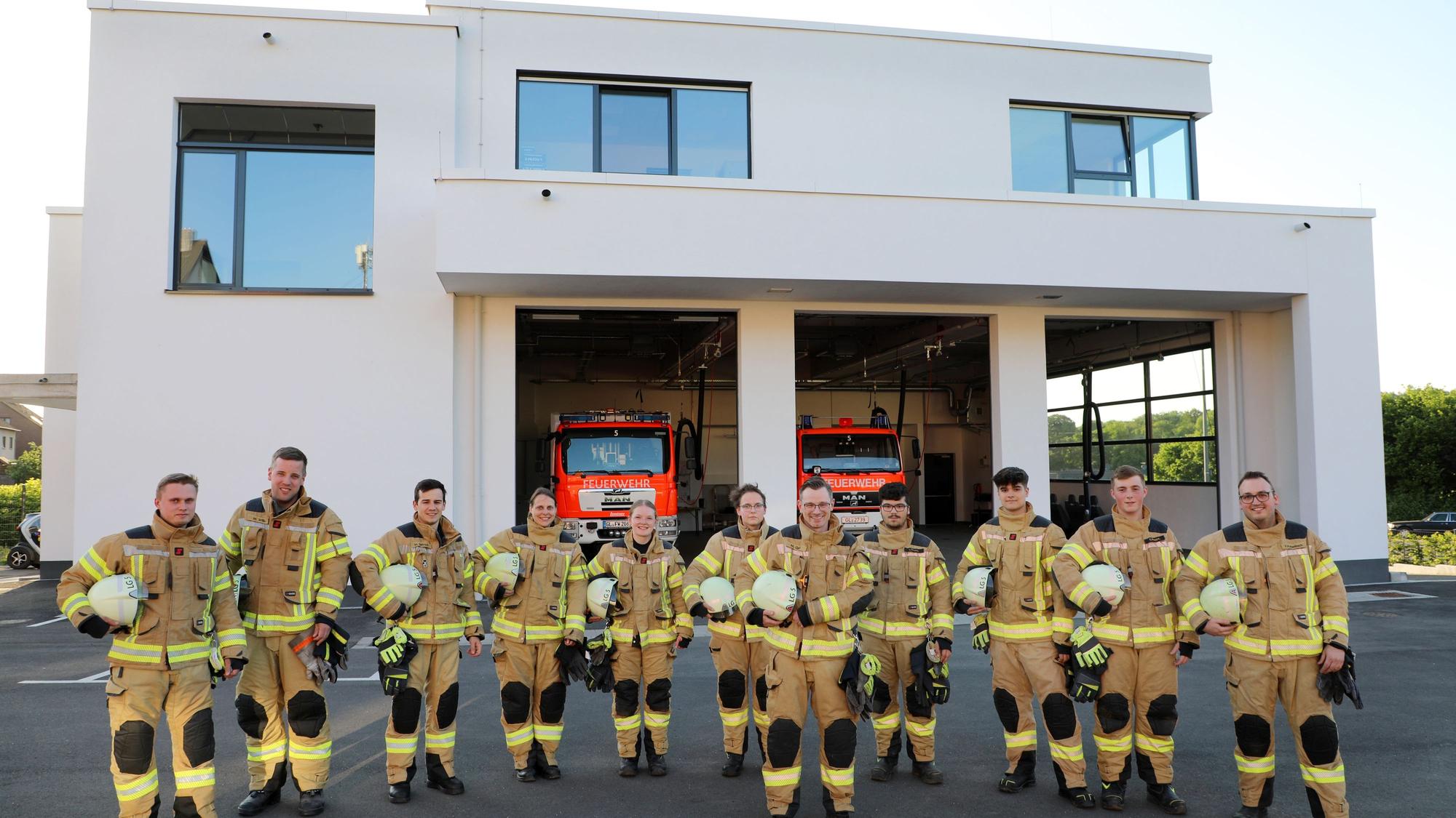 Feuerwehrleute stehen vor einem Feuerwehrhaus mit geöffneten Fahrzeughallentoren, in denen zwei große Einsatzfahrzeugezu sehen sind.