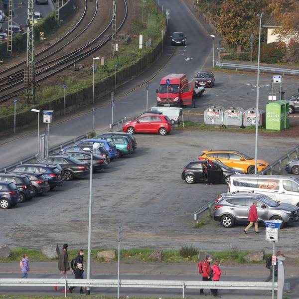 Parkende Autos stehen auf einer Schotterfläche. Links am Bildrand verläuft die rechtsrheinische Bahntrasse.