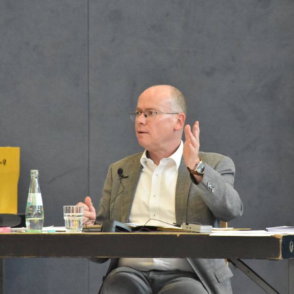 Bürgermeister Mario Loskill gestikuliert mit erhobener Hand und sitzt an einem Schreibtisch in einer Sporthalle.