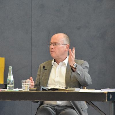 Bürgermeister Mario Loskill gestikuliert mit erhobener Hand und sitzt an einem Schreibtisch in einer Sporthalle.