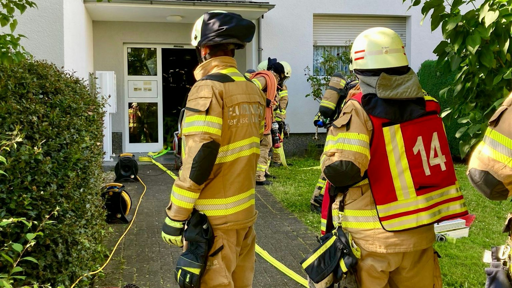 Feuerwehrfleute stehen vor dem Eingang eines Mehrfamilienahuses. Ein Feuerwehrschlauch liegt ausgerollt auf dem Boden und führt in den Hauseingang.
