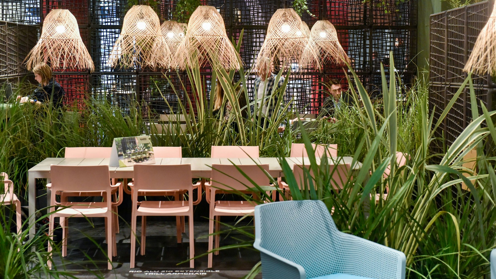 Auf einer Messe sind ein Sessel, Tisch und darüberhängede Lampen in künstlichem, hohen Gras ausgestellt.