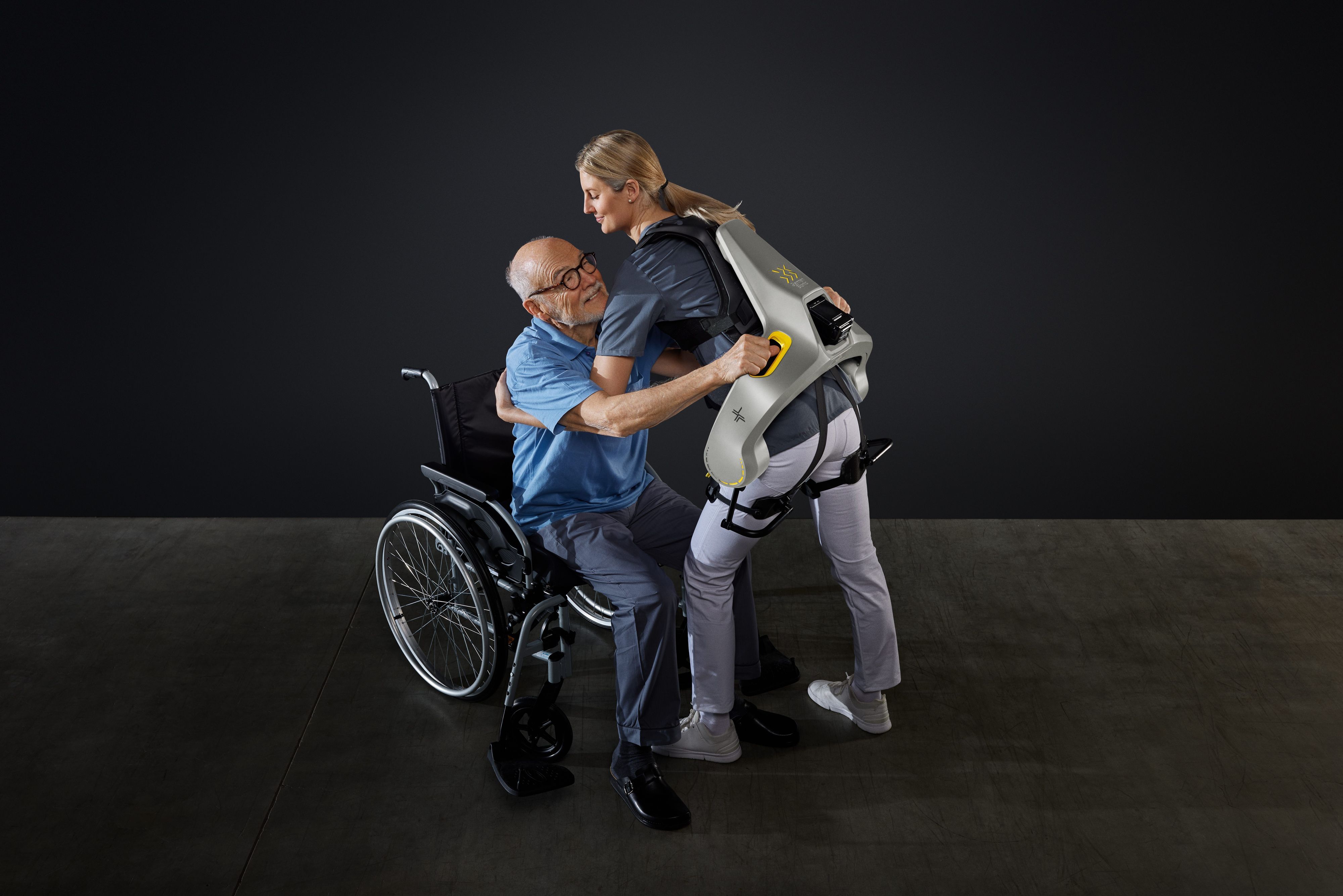 Eine Frau hebt mit Hilfe eines intelligenten Kraftanzugs der Firma German Bionic einen Mann aus einem Rollstuhl.