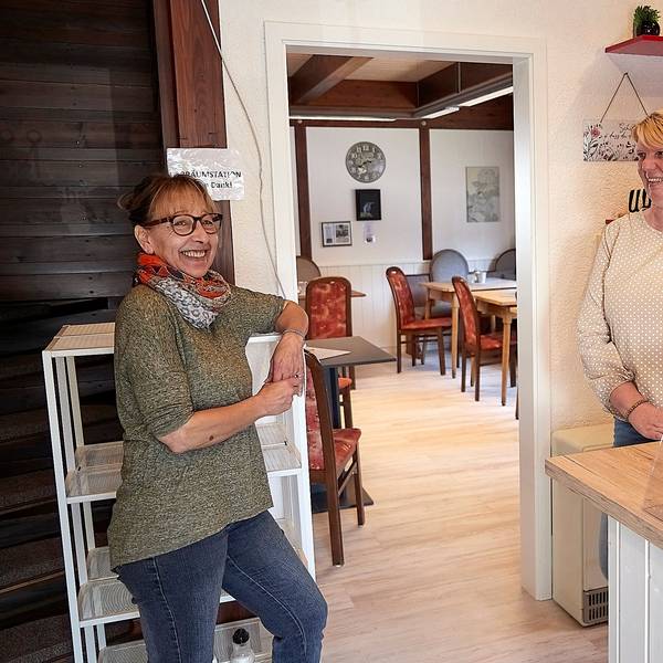 Das Bild zeigt zwei Frauen, die in einem Café an einem Regal und einer Theke stehen.
