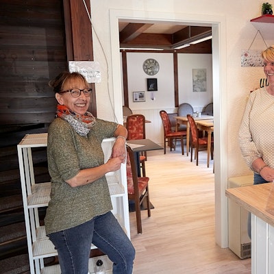 Das Bild zeigt zwei Frauen, die in einem Café an einem Regal und einer Theke stehen.