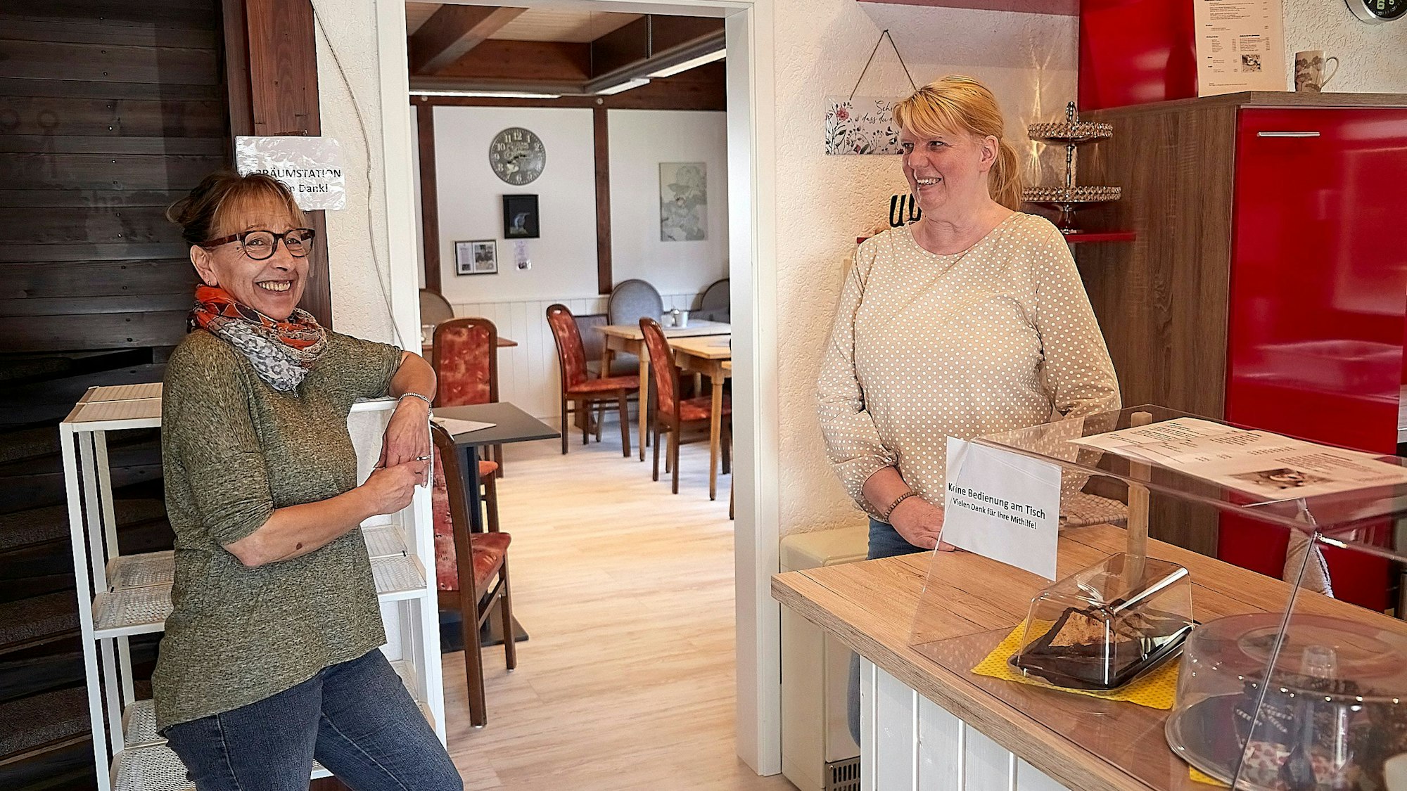 Das Bild zeigt zwei Frauen, die in einem Café an einem Regal und einer Theke stehen.