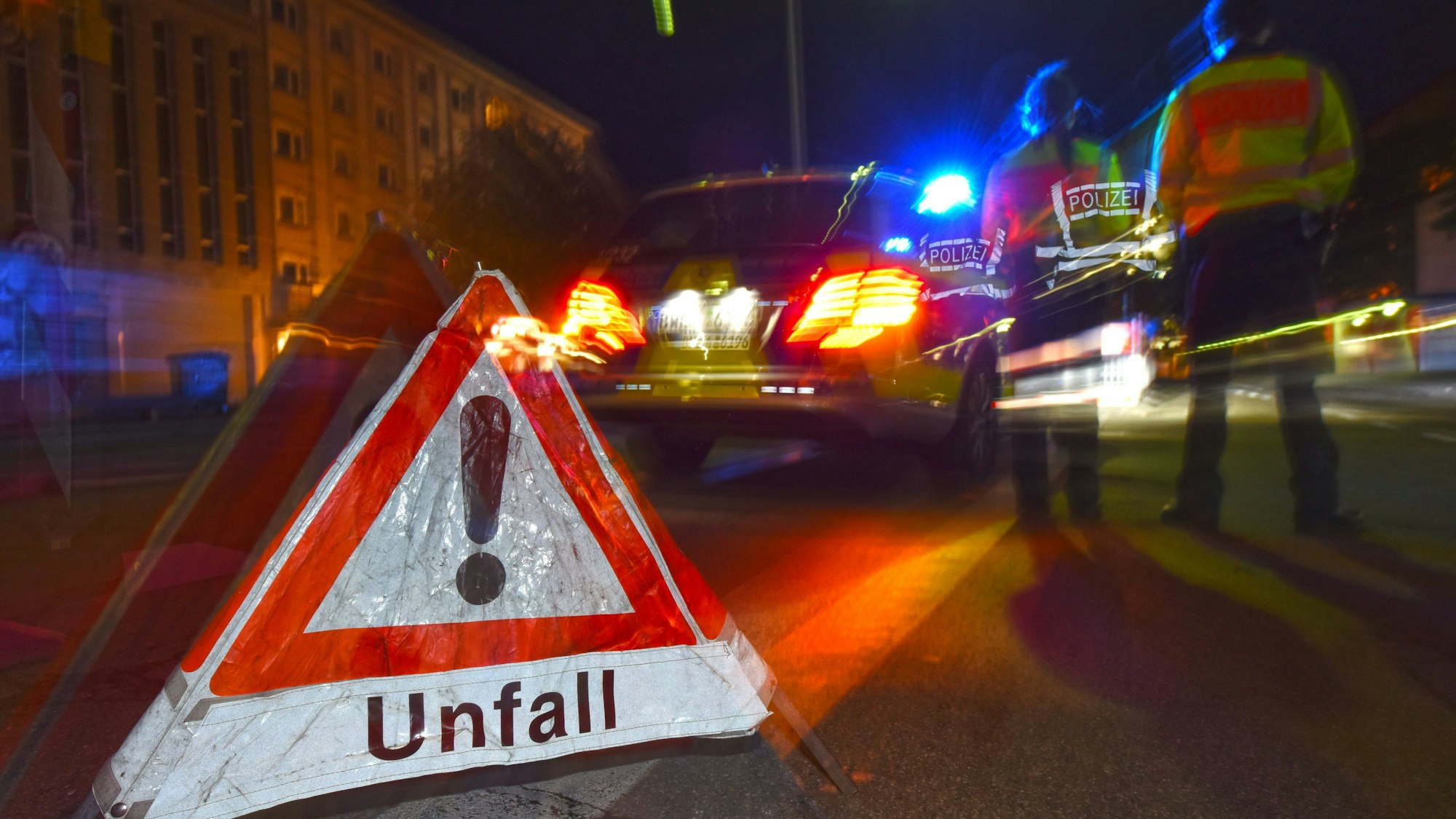 Ein Polizeifahrzeug steht mit Blaulicht auf der Straße. Daneben steht ein Warndreieck mit dem Schriftzug „Unfall“.