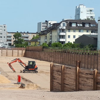 Im Hintergrund die Bebauung entlang der Neusser Straße entlang der Baugrube bei gutem Wetter. In der Baugrube steht ein kleiner Bagger.