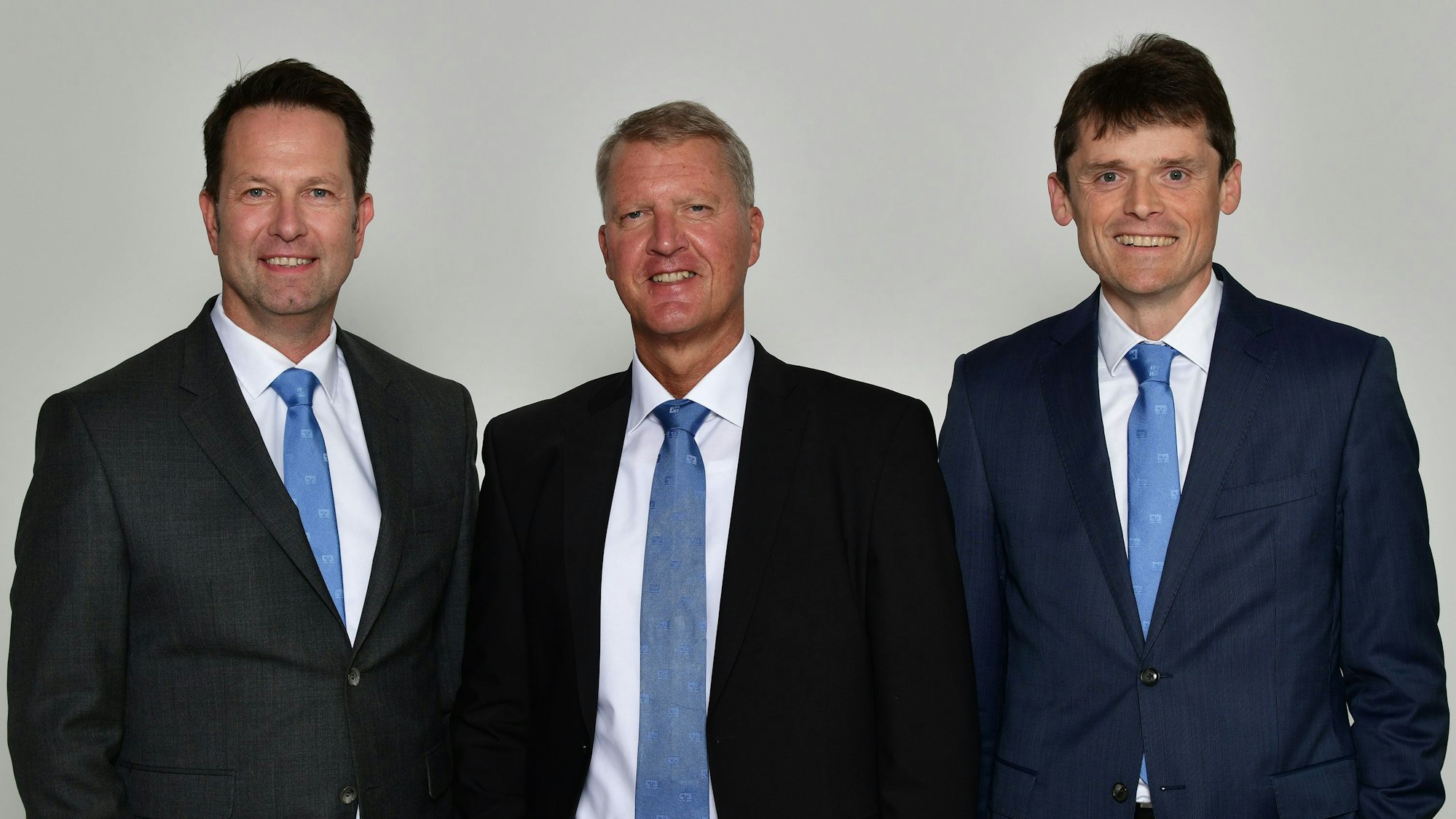 Drei Männer in Anzügen und mit blauen Krawatten stehen nebeneinander und schauen in die Kamera.