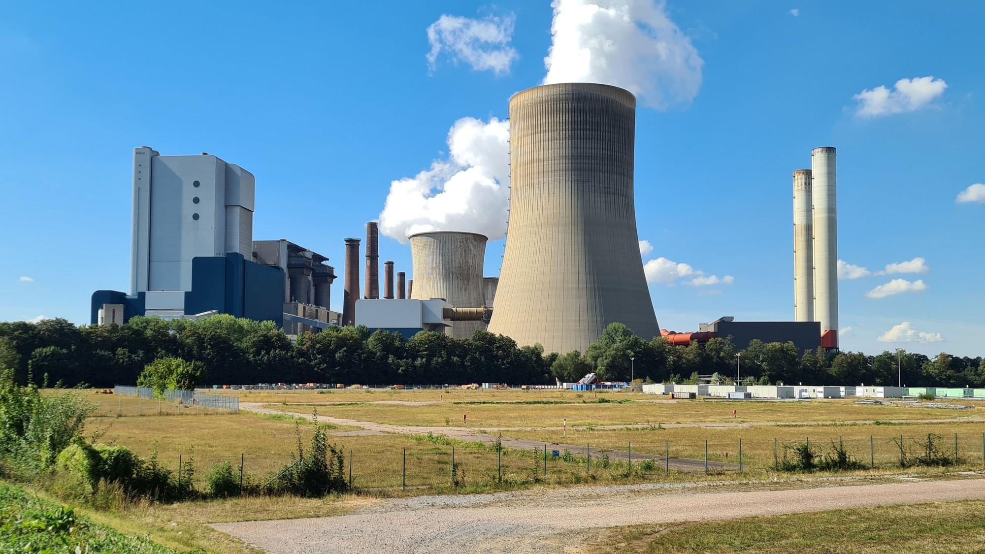 Hinter einer brachliegenden Wiese erhebt sich ein Kraftwerk mit Kühltürmen, aus denen weiße Schwaden aufsteigen.