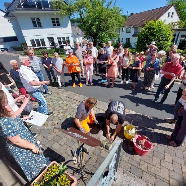 Zwei Männer verlegen kniend Stolpersteine im Pflaster, eine Gruppe von Menschen beobachtet und fotografiert die Aktion.