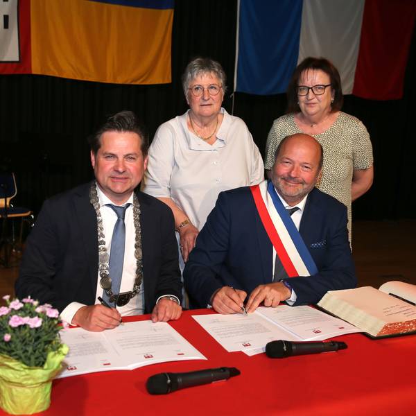 Zwei Bürgermeister sitzen an einem Tisch mit roter Tischdecke und unterzeichnen einen Städtepartnerschaftsvertrag. Daneben liegt das Goldene Buch der Stadt Wiehl. Im Hintergrund stehen zu Frauen.