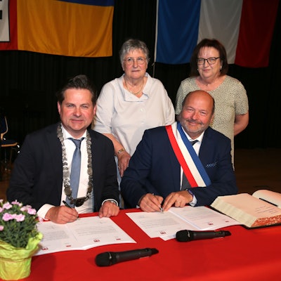 Zwei Bürgermeister sitzen an einem Tisch mit roter Tischdecke und unterzeichnen einen Städtepartnerschaftsvertrag. Daneben liegt das Goldene Buch der Stadt Wiehl. Im Hintergrund stehen zu Frauen.