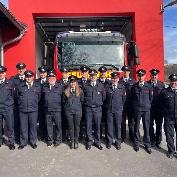 16 Feuerwehrmänner und -frauen stehen vor dem neuen Anbau einer Löschwache. In einer Garage steht ein großes Feuerwehrauto.