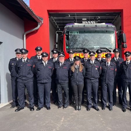 16 Feuerwehrmänner und -frauen stehen vor dem neuen Anbau einer Löschwache. In einer Garage steht ein großes Feuerwehrauto.
