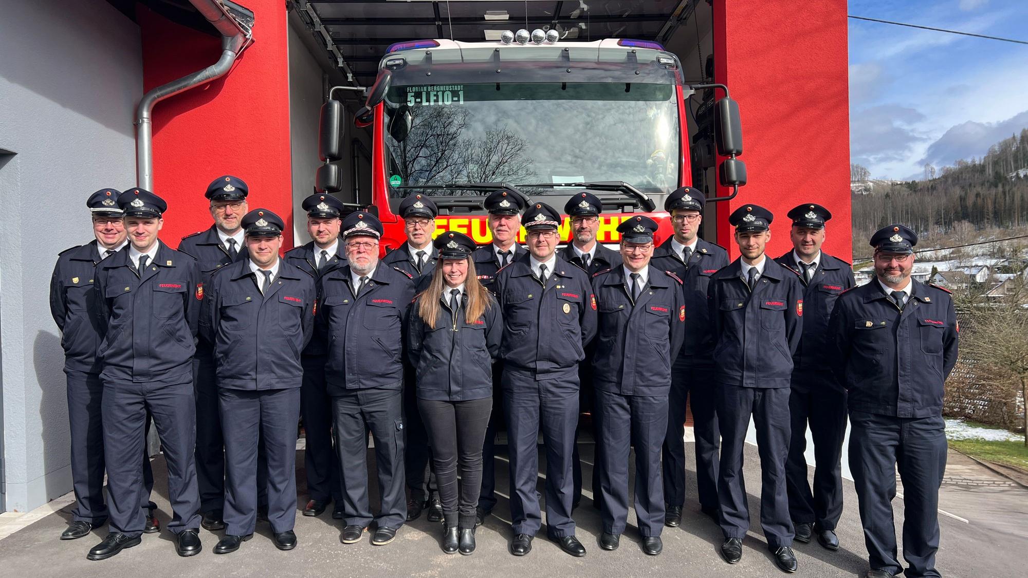 16 Feuerwehrmänner und -frauen stehen vor dem neuen Anbau einer Löschwache. In einer Garage steht ein großes Feuerwehrauto.