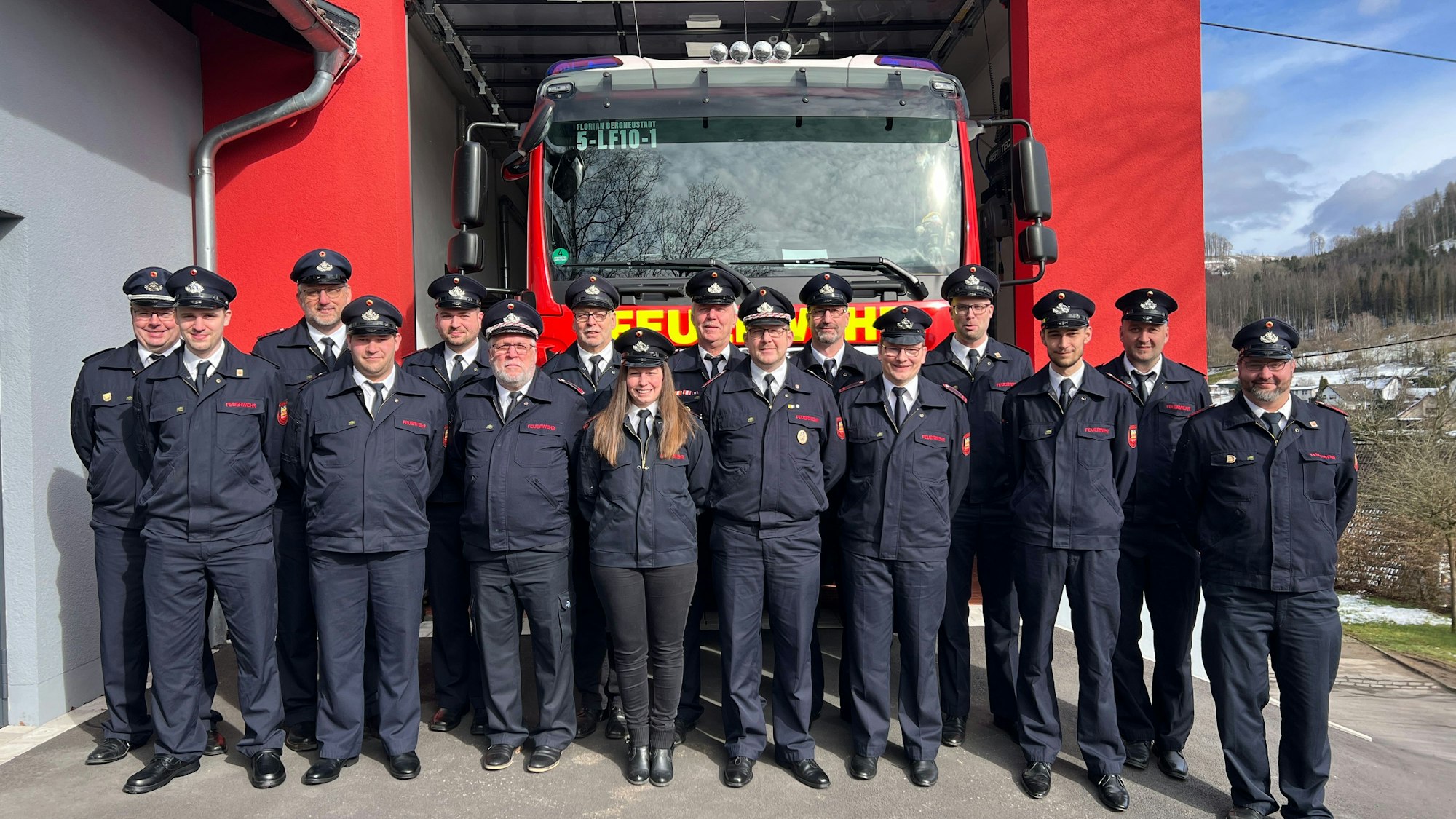 16 Feuerwehrmänner und -frauen stehen vor dem neuen Anbau einer Löschwache. In einer Garage steht ein großes Feuerwehrauto.
