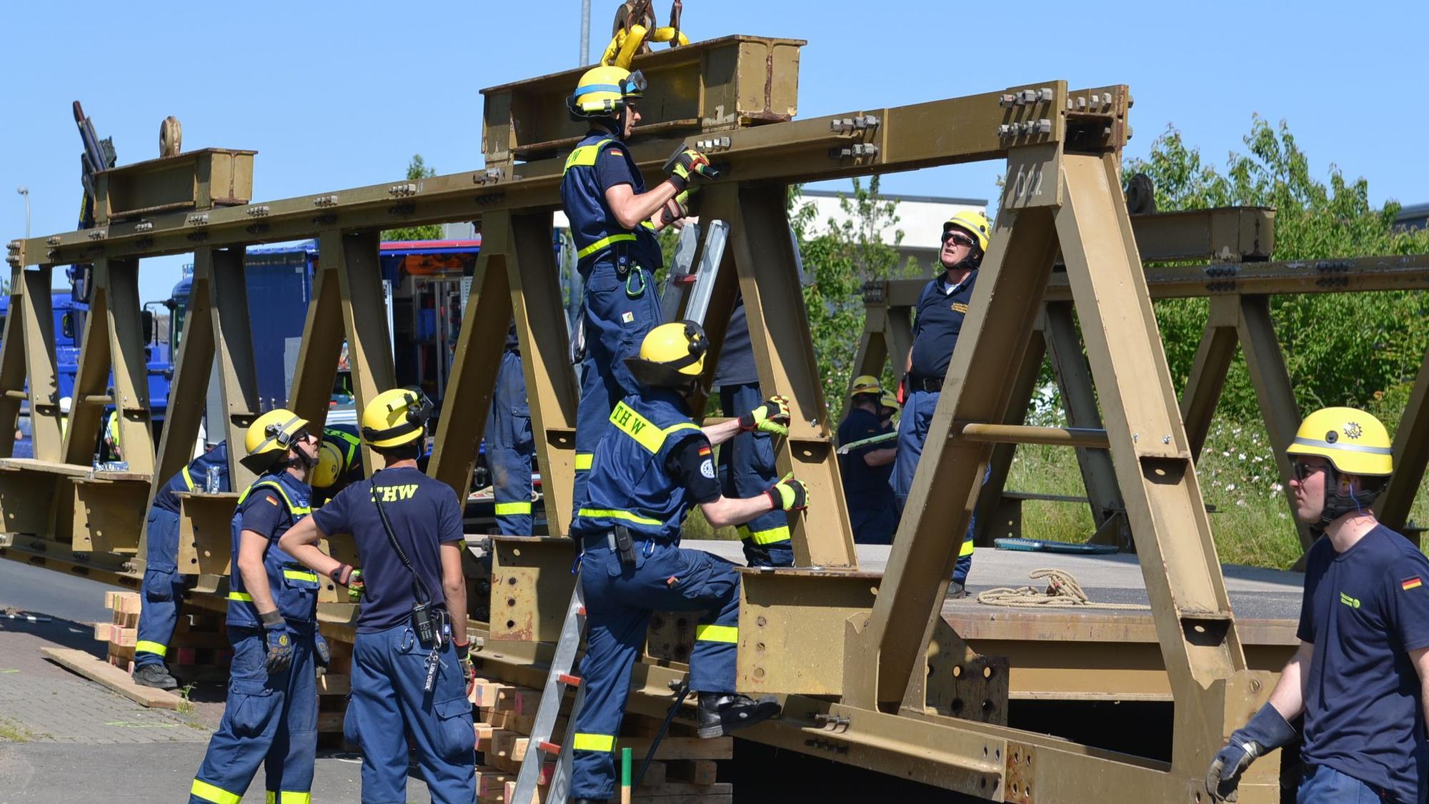 Acht THW-Helfer in blauer Arbeitskleidung und mit gelben Helmen arbeiten am Abbau einer massiven Stahlbrücke.