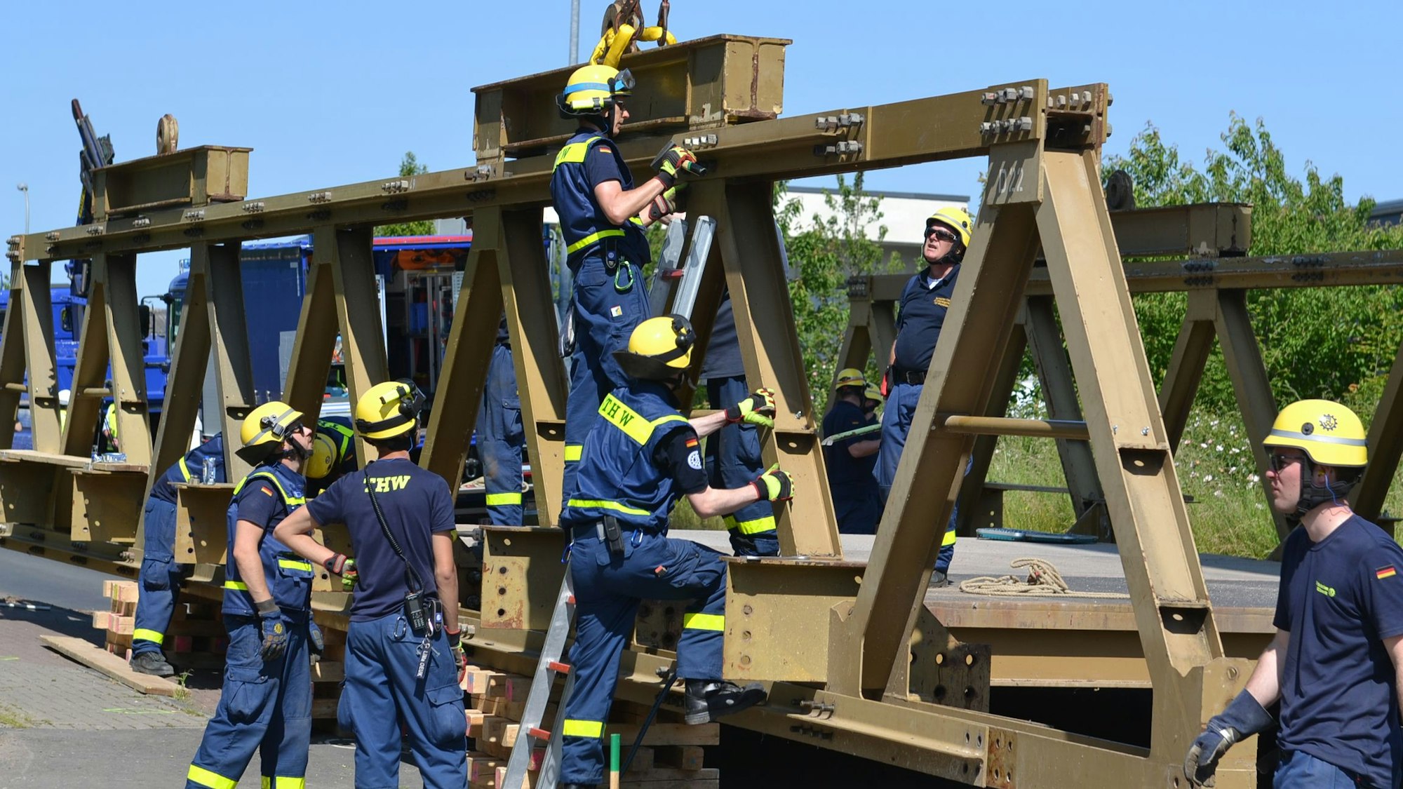 Acht THW-Helfer in blauer Arbeitskleidung und mit gelben Helmen arbeiten am Abbau einer massiven Stahlbrücke.