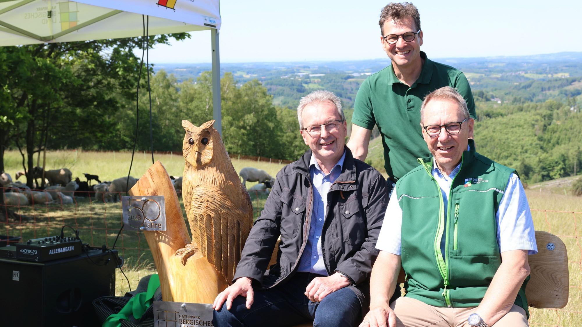 Drei Männer sitzen und stehen an einer Bank aus Holz, an deren Seite eine geschnitzte Eule zu sehen ist.