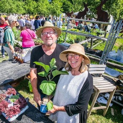 Ein Mann und eine Frau mit Hüten zeigen fröhlich eine Topfpflanze an einem Stand.