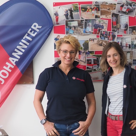 Zwei Frauen stehen vor einer Wand mit Fotos neben einem „Johanniter“-Banner.